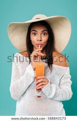Image of cheerful young woman wearing hat posing over blue background while drinking juice.