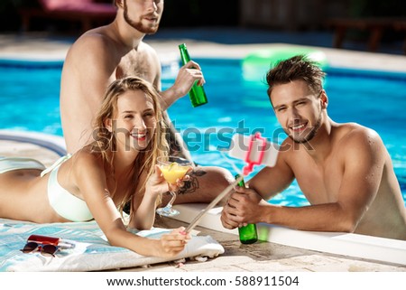 Friends smiling, making selfie, drinking cocktails, relaxing near swimming pool.