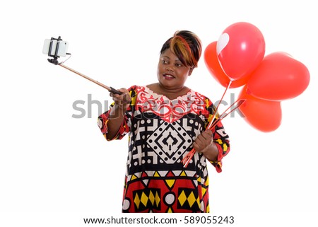 Happy fat black African woman smiling while holding bunch of red balloons with heart sign and taking selfie picture with selfie stick