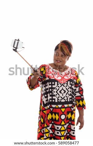 Studio shot of happy fat black African woman smiling while holding selfie stick and taking selfie picture with mobile phone