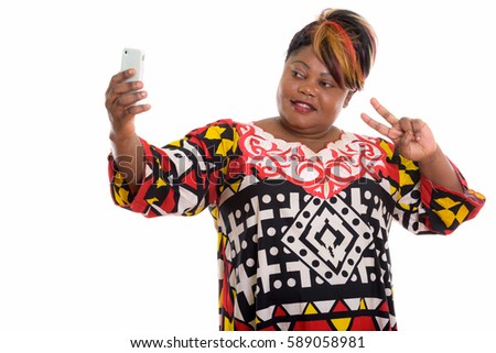 Studio shot of happy fat black African woman smiling while taking selfie picture with mobile phone and giving peace sign