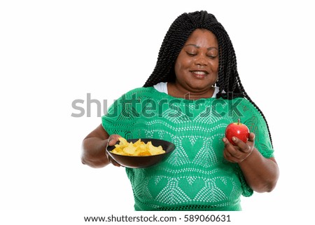 Studio shot of happy fat black African woman smiling while holding bowl of potato chips and looking at red apple