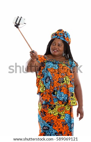 Happy fat black African woman smiling while holding selfie stick and taking selfie picture with mobile phone while wearing traditional clothes