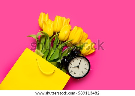 bunch of beautiful yellow tulips in cool yellow shopping bag and black alarm clock on the wonderful pink background