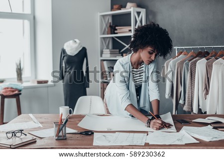 Turning ideas into clothing. Beautiful young African woman working on sketches while standing in her studio near the clothes hanging on the racks