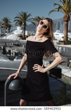 Fashion portrait of luxury blonde woman posing near yacht club in black stylish dress and sunglasses.