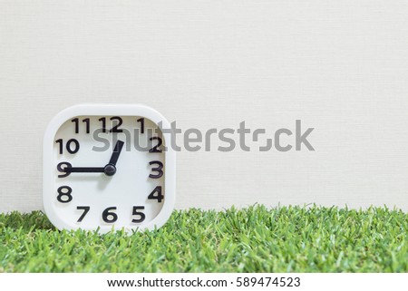 Closeup white clock for decorate show a quarter to one o'clock or 12:45 a.m. on green artificial grass floor and cream wallpaper textured background with copy space