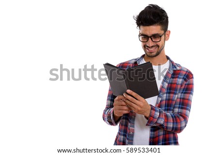 Studio shot of young happy Persian man smiling while wearing eyeglasses and reading book
