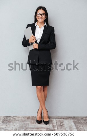 Full length portrait of a happy smiling business woman holding laptop and looking at camera over gray background