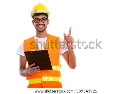 Studio shot of young happy Persian man construction worker smiling and holding clipboard while pointing finger up