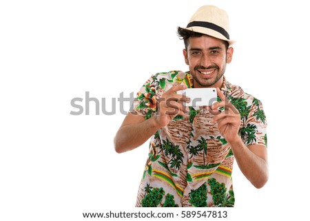 Studio shot of young happy Persian tourist man smiling while taking picture with mobile phone and looking at camera