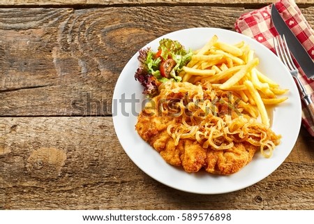 Schnitzel served with fried onion, salad and french-fries on white plate, with cutlery over red napkin from above shot on rustic rough wooden table surface with copy space