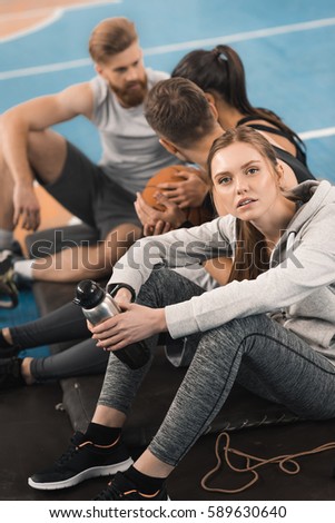 Young sporty woman holding bottle while sitting near teammates and looking at camera
