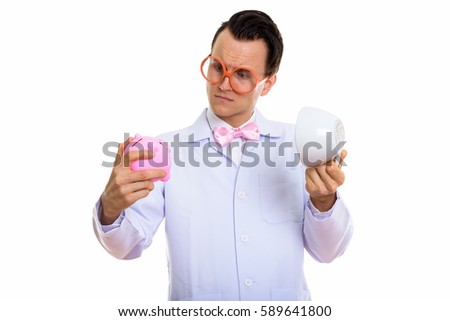 Studio shot of young crazy man doctor looking at piggy bank while holding empty coffee cup