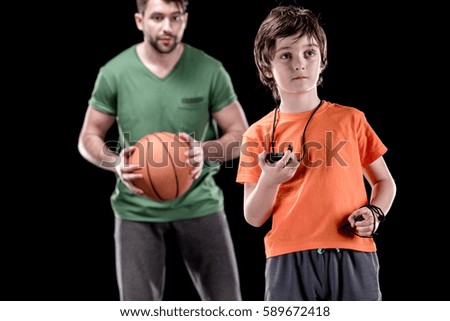 boy with stopwatch controlling time with man holding basketball ball isolated on black, focus on foreground