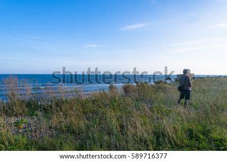 Man holding a selfie stick standing on tall grass by a calm blue sea in summer