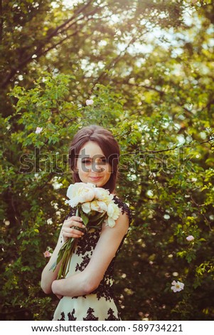 Girl with bouquet of peonies stands before green bush with tiny white flowers