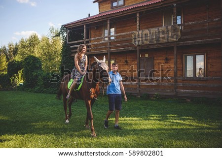Romantic young couple in love, a walk on a horse on nature background and wooden country-style hotel. Young woman sitting on horseback. A man walks around, holds . Concept: love, romance, Hobbies.