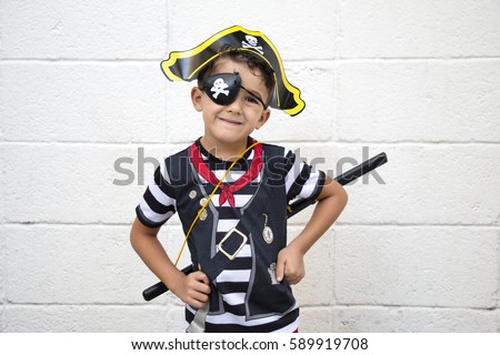 Little pirate boy is posing to the camera while his hands on waist. Rustic wall background.