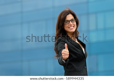 Professional businesswoman doing thumbs up gesture against corporate building outside. Female business success and job goals.