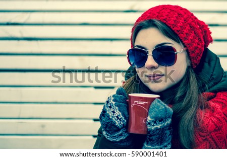 Portrait of a beautiful young girl in sunglasses and red hat with a scarf, which is holding a red cup of hot tea and sitting on a park bench