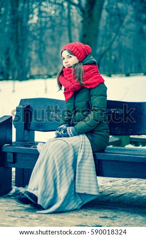 Portrait of a beautiful young girl in a red cap with a scarf, which sits under a blanket on a bench in the park in winter