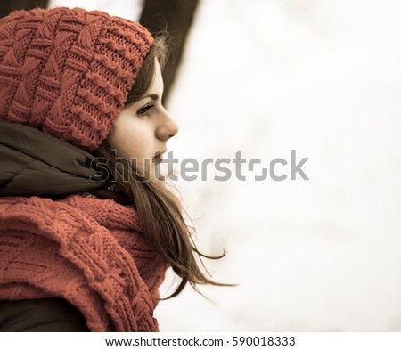 Portrait of a beautiful young girl in a red cap with a scarf, which sits under a blanket on a bench in the park in winter