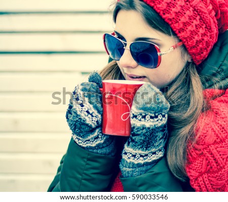 Portrait of a beautiful young girl in sunglasses and red hat with a scarf, which is holding a red cup of hot tea and sitting on a park bench