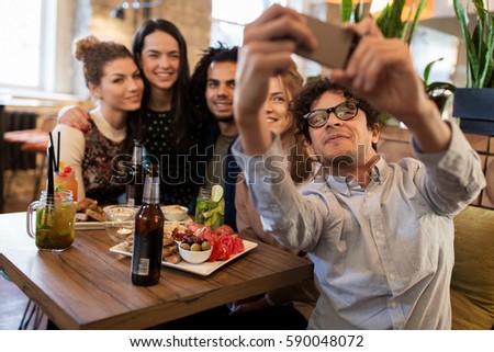 leisure, technology, friendship, people and holidays concept - happy friends with food and drinks taking selfie by smartphone at bar or cafe