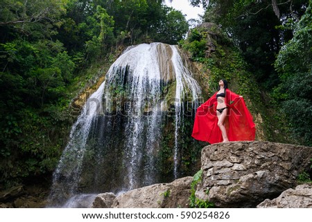 Young pretty brunette woman in red long dress is standing near beautiful waterfall in hot tropical jungle forest at summer sunny day