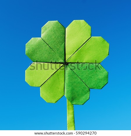 Green paper origami folded shamrock on blue sky background. Sunny weather outdoors.