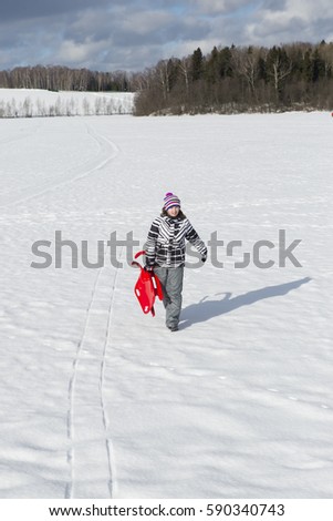 Girl with red sled in the hands walking on snow-covered field