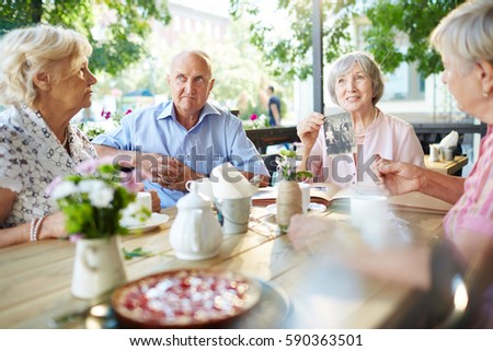 Elderly woman in pastel pink blouse and pearl necklace sharing story of her family and showing black-and-white photos to her friends in cafe