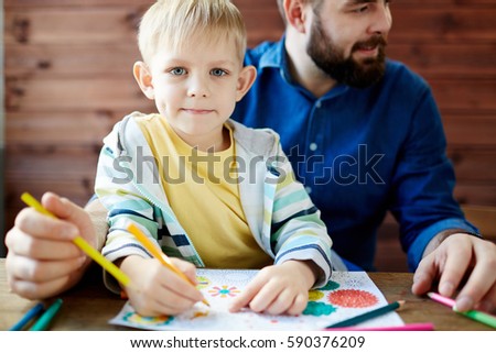Cute little blond-haired boy with deep grey eyes sitting on laps of his bearded father, looking at camera and holding pencil in hand for coloring picture
