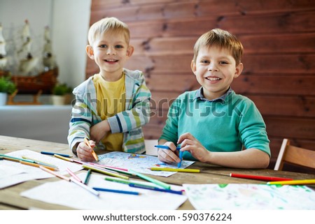 Two funny little brothers with wide smiles distracted for a second from drawing colorful picture in order to pose for photography in art class