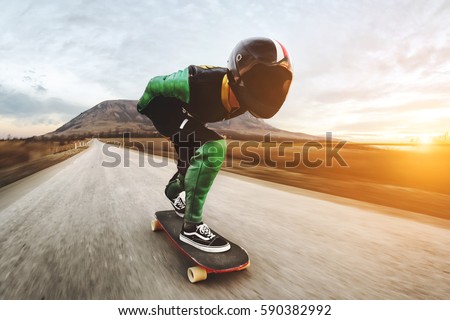 A young man in helmet and a leather suit in a special rack rides a longboard on afsaltu on background mountains and beautiful sky Royalty-Free Stock Photo #590382992