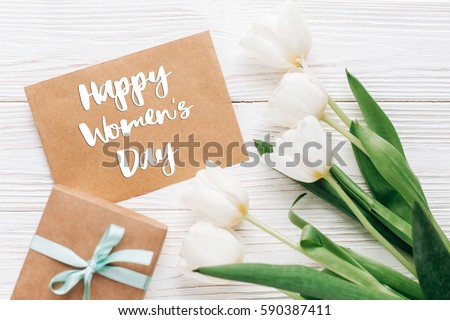 happy womens day text sign on stylish craft present with greeting card and tulips on white wooden rustic background. flat lay with flowers and gift with space for text. greeting card