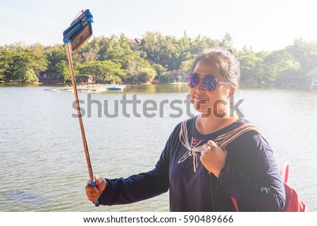 Asian woman is enjoy taking selfie picture by her own mobile phone and selfie stick in the park somewhere with the nice flare in the afternoon.