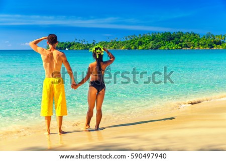 A couple in love, holding hands enjoying the vacation in tropical paradise island with green palms, sandy beach and clean sea ocean water on a background colorful blue sky
