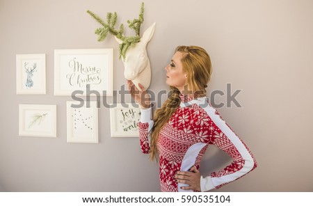 beautiful blonde girl standing against a background of gray wall with white head of decorative deer with horn. Christmas and New Year concept. red knitted costume. white picture frame hang on wall. 