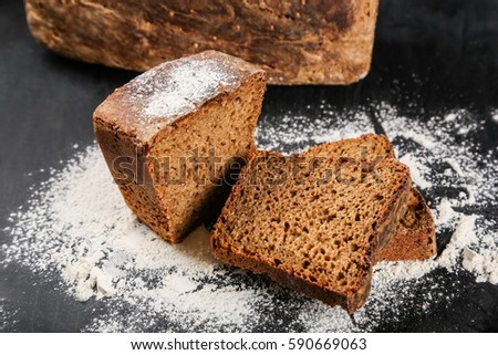 On pictures cut homemade rye bread on the black table, top view, largely cut into pieces, sprinkled with white meal, in the background blurred a whole loaf of bread