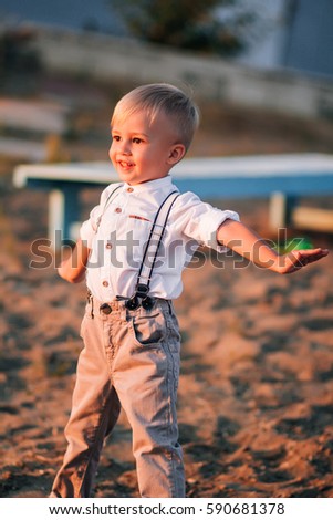 Beautiful boy with white hair, stylish clothes and a charming smile. A child on the sandy shore. Playing near the pond. Stylish baby pictures