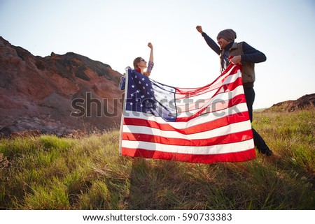 Couple of joyous young people waving big American banner jumping on top of hill in mountains