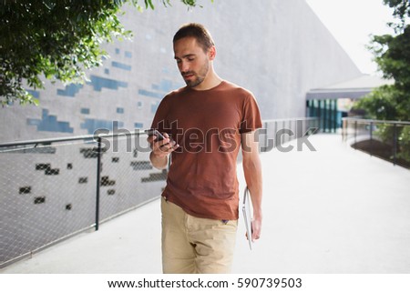 Young bearded man in casual clothes going along the path and texting message on his smartphone