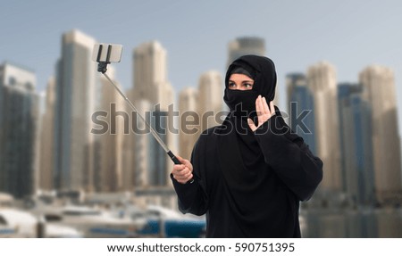 technology and people concept - muslim woman in hijab taking picture with smartphone selfie stick over dubai city street background