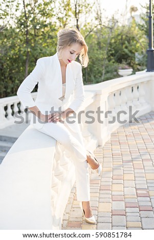 Business young blonde woman in a white pantsuit with red lips sits on a colonnade against the backdrop of the blue sea in the evening at sunset. Business and partnerships, women's rights in business