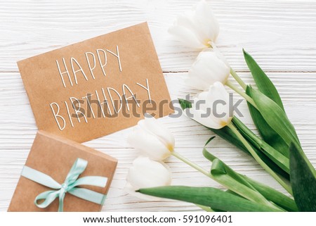 happy birthday text sign on stylish craft present with greeting card and tulips on white wooden rustic background. flat lay with flowers and gift with space for text.  greeting card