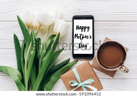 happy mothers day text sign on stylish gift and phone and tulips and coffee on white wooden rustic background. flat lay with flowers and gadget with empty screen with space for text.