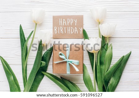 happy birthday text sign on greeting card with stylish present box and tulips on white wooden rustic background. flat lay mock up with flowers and empty paper with space for text