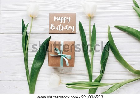 happy birthday text sign on mock up greeting card with stylish present box and tulips on white wooden rustic background. flat lay with flowers and empty paper with space for text.